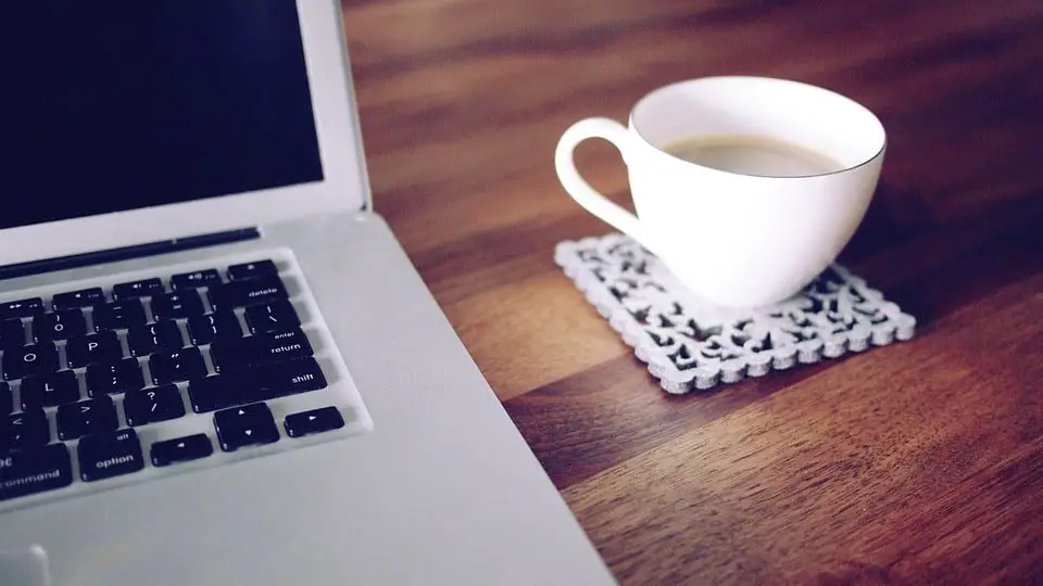 A coffee/tea cup placed on an artistic toaster next to a laptop on a wooden table.